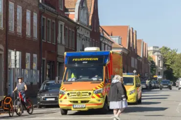 Rettungswagen der Feuerwehr fährt durch eine Straße mit Backsteinhäusern, daneben Radfahrer