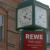 Straßenuhr über einem REWE-Schild mit Adresse und Öffnungszeiten vor Altbauten