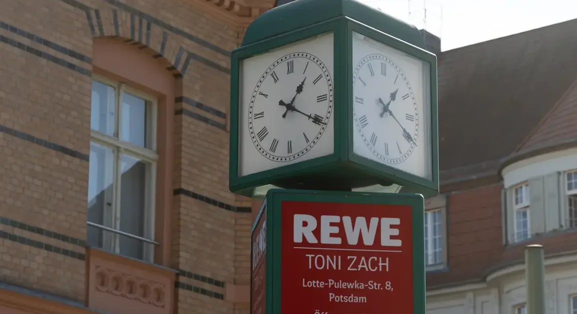 Straßenuhr über einem REWE-Schild mit Adresse und Öffnungszeiten vor Altbauten