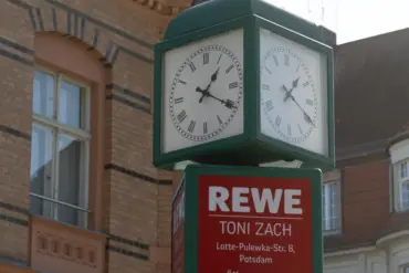 Straßenuhr über einem REWE-Schild mit Adresse und Öffnungszeiten vor Altbauten