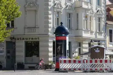 Litfaßsäule und Baustellenabsperrung vor einem Altbau mit Restaurant