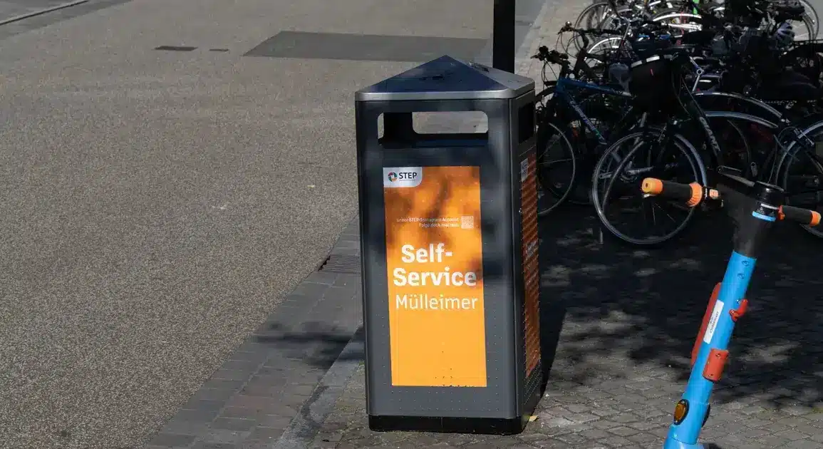 Orangefarbener Self-Service-Mülleimer der Stadtreinigung, dahinter abgestellte Fahrräder
