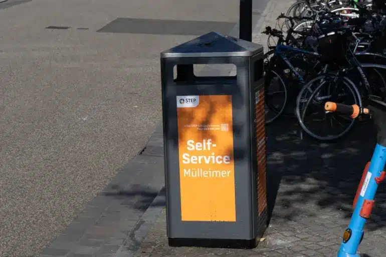 Orangefarbener Self-Service-Mülleimer der Stadtreinigung, dahinter abgestellte Fahrräder